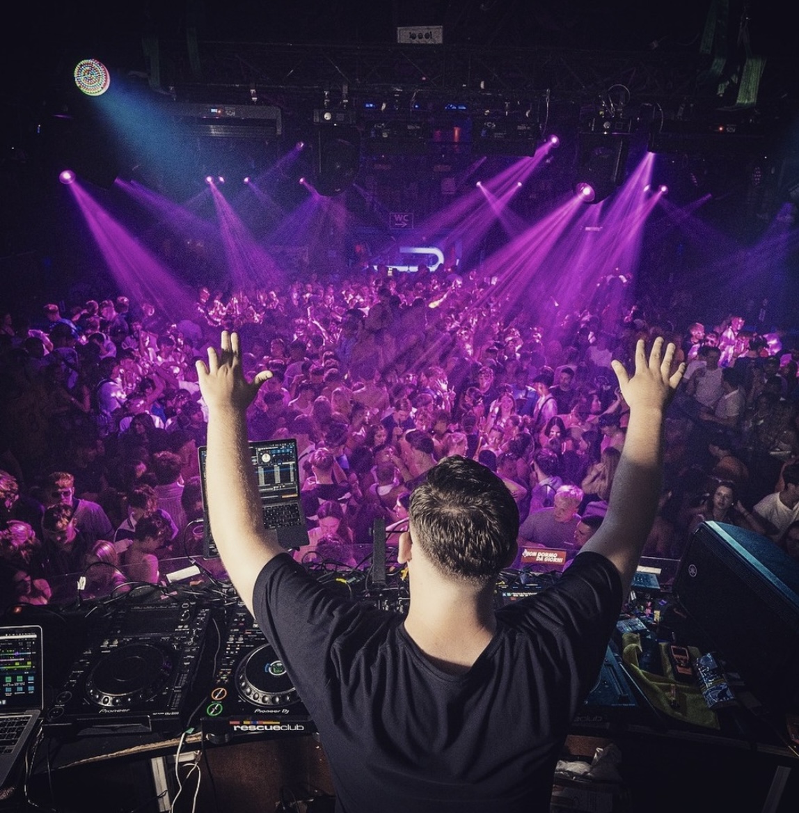 Vextra behind the decks with hands raised over a packed club floor
